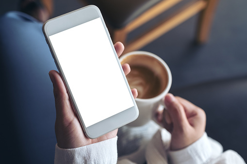 Top view mockup image of woman's hands holding white mobile phone with blank screen while drinking coffee in cafe