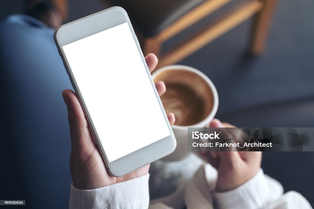 Top view mockup image of woman's hands holding white mobile phone with blank screen while drinking coffee in cafe Telephone Stock Photo