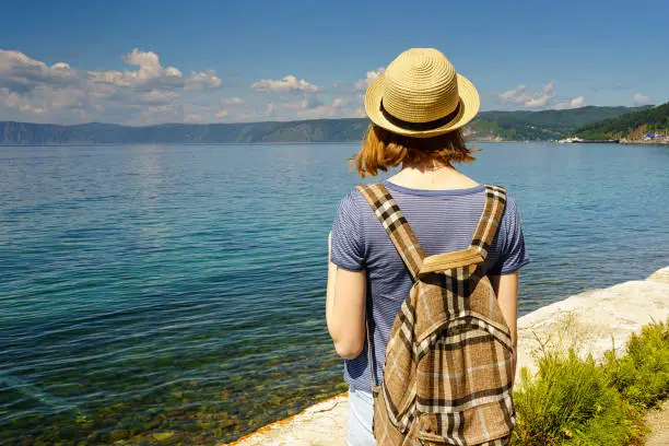 Young lady with backpack admiring the summer landscape of lake Baikal from the waterfront of the Listvyanka village