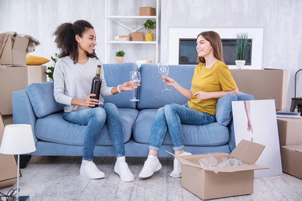 Charming young girls celebrating moving into new flat Mini celebration. Upbeat young girls sitting on the sofa being about to drink some wine while celebrating moving in to a new apartment college dorm party stock pictures, royalty-free photos & images