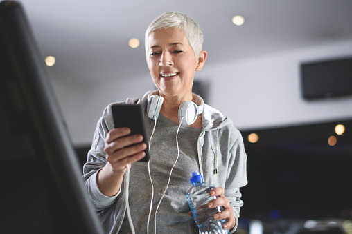 Active senior female with white headphones exercising on the treadmill in the health club and using mobile phone.