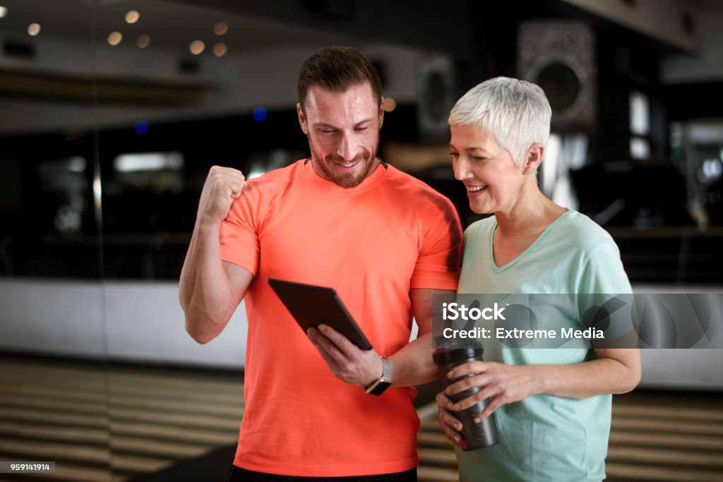 Active senior woman and fitness instructor Active senior female in the health club being motivated by personal trainer with multimedia content on digital tablet. Coach Stock Photo