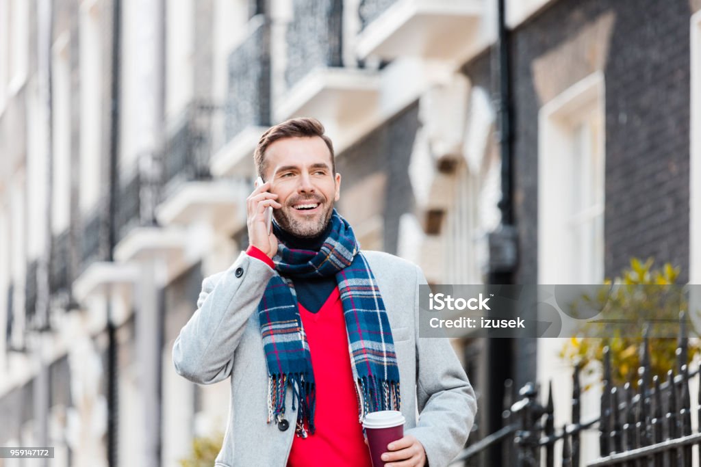 Elegant smiling man talking on phone on the London street Handsome man wearing coat talking smart phone on the London street, holding cup of coffee. Autumn season. Adult Stock Photo