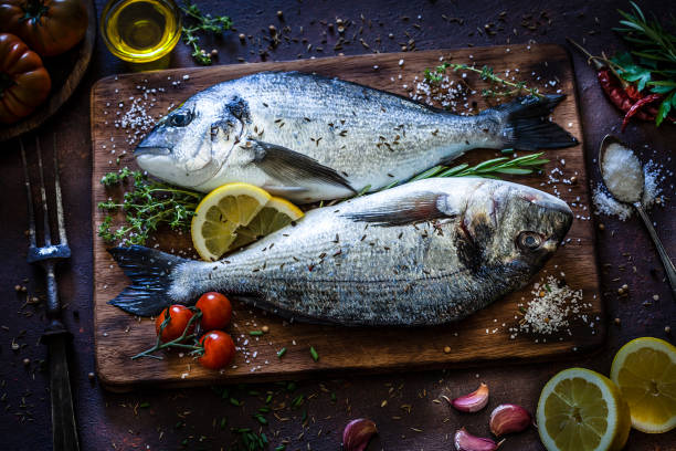 Top view of two fresh sea bream on a wooden cutting board ready for cooking with herb and spices shot on rustic kitchen table. The pan is surrounded by many ingredients and spices for cooking fish like lemon, garlic, tomatoes, parsley, chive, olive oil, salt, pepper and others. Low key DSRL studio photo taken with Canon EOS 5D Mk II and Canon EF 100mm f/2.8L Macro IS USM