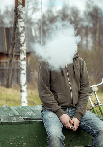 Millennial man in a casual clothing is resting sitting on crate next to retro bike. He is smoking the electronic cigarette. The man's head is not visible beyond a smoke cloud. Shooting in a countryside at spring day