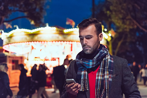 Outdoor portrait of handsome man wearing winter clothes using smart phone in front of illuminated carousel in London.