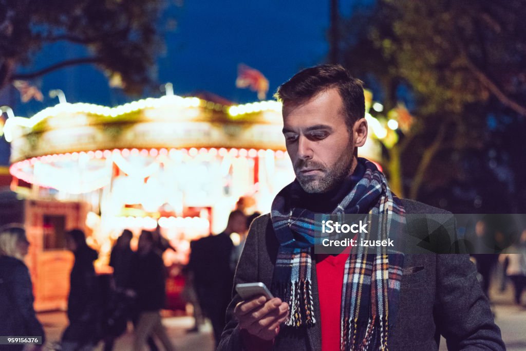 Handsome man using smart phone in front of illuminated carousel Outdoor portrait of handsome man wearing winter clothes using smart phone in front of illuminated carousel in London. Carousel Stock Photo