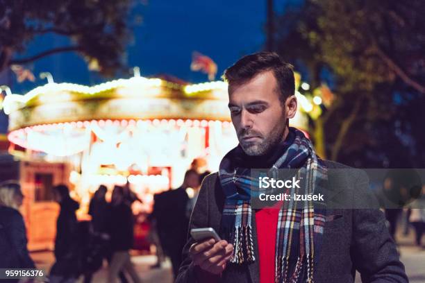 Handsome Man Using Smart Phone In Front Of Illuminated Carousel Stock Photo - Download Image Now