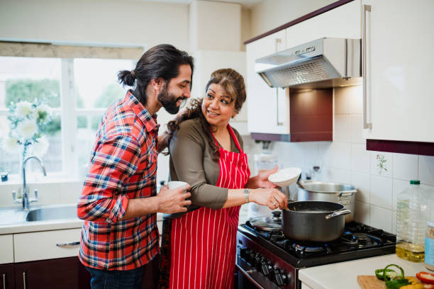 Do You Need me to Taste Test it, Mum? Mature woman is cooking in her kitchen and her son is looking over her shoulder to see what she is making. adult offspring stock pictures, royalty-free photos & images