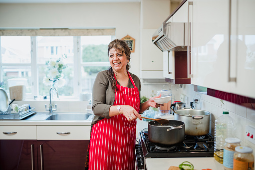 Mature woman is talking and laughing with someone out of the frame while she makes dinner at the cooker.