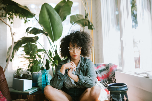 A woman relaxes in her house, eyes closed in prayer, deep thought, or meditation.  A healthy part of living with mindfulness.