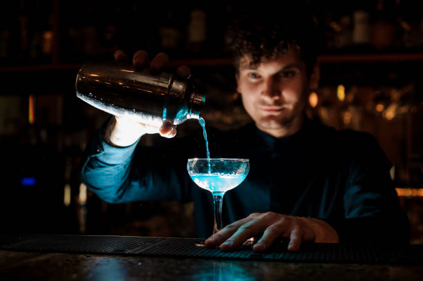 istock Young smiling barman pouring fresh drink with blue liquor from a shaker into a glass using strainer 959252230