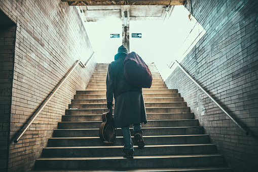 One man, young homeless man holding acoustic guitar, walking up the stairs, rear view.