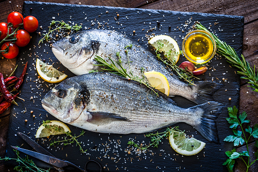 Top view of two fresh sea bream on a dark slate board ready for cooking with herb and spices shot on rustic kitchen table. The pan is surrounded by many ingredients and spices for cooking fish like lemon, garlic, tomatoes, parsley, chive, olive oil, salt, pepper and others. Low key DSRL studio photo taken with Canon EOS 5D Mk II and Canon EF 100mm f/2.8L Macro IS USM