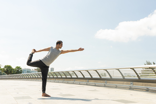 Indian man doing lord of dance pose outdoors on bridge on sunny day. City yoga concept. Side view.