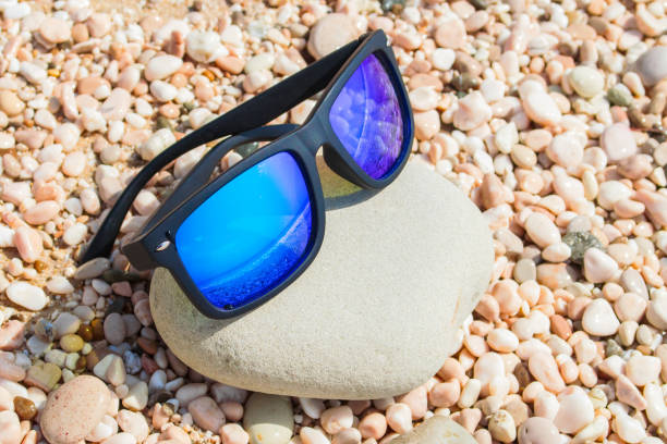 istock Blue sunglasses on a pebble beach reflecting the ocean. Summer holidays concept 959352408