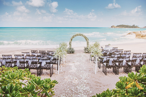 Beach wedding ceremony venue setting on the beach, white sand with panoramic ocean background, tree in foreground, clear sky, flowers floral decoration for arch, the roses petal on the aisle walk way