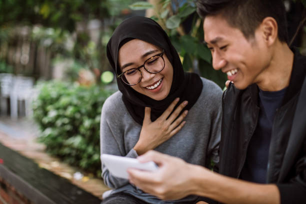 Charmed By Things He's Shown Her On Phone Young Multi-ethnic Couple Went Out On a Date Outdoors. Guy Is Showing His Girlfriend Interesting Things On Internet And She Seems To Be Amazed By It indonesian ethnicity stock pictures, royalty-free photos & images