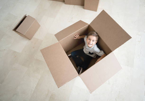 A handsome boy proudly holds a box full of his old toys, ready for donation. The boy donates his toys packed in a cardboard box, sits in the living room, looks at the camera.