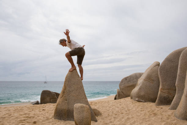 mature man scrambles onto top of rock triangle on beach - balance simplicity nature beach imagens e fotografias de stock