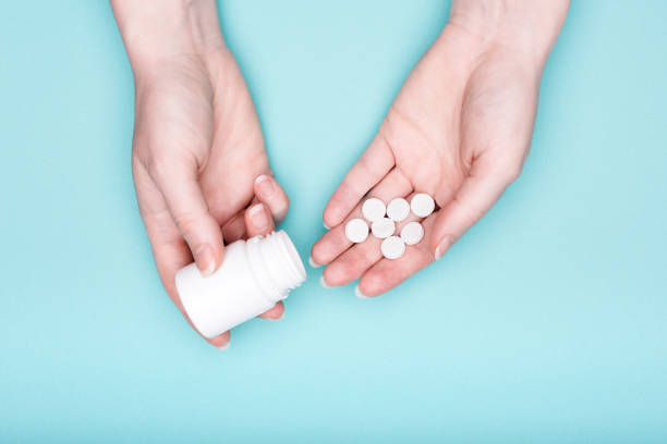 close up of female hands holding medication bottle and white pills over pastel blue background. patient taking medication. - bottle vitamin pill nutritional supplement white imagens e fotografias de stock