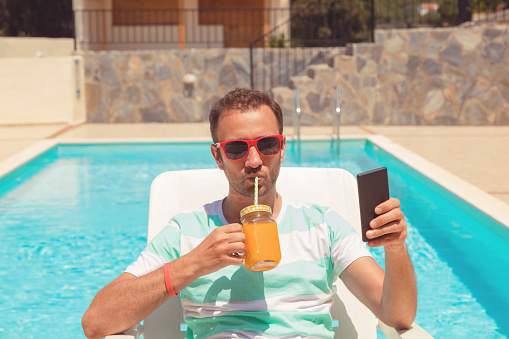 Modern man using cellphone while drinking juice on the pool.
