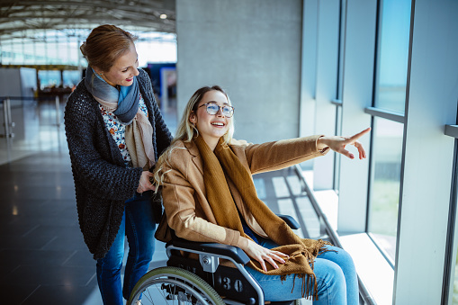 Young disabled woman on wheelchair and carer waiting at airport and looking at window view