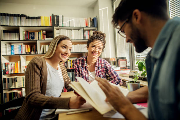 istock Two young joyful female students are smiling at and taking notes from a male lecturer while sitting at the desk near window and bookshelf in the school library. 959533726