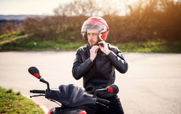Drive safe! Young handsome biker man in black leather jacket put on a red helmet while sitting on a scooter.