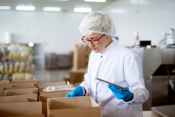istock Young gorgeous concerned female worker in sterile cloths using a tablet to check correction of inventory within boxes in factory storage room. 959555276