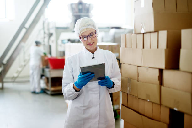 istock Young female dedicated worker is checking her tablet while standing near the big stack of cardboard boxes in the factory. 959556220