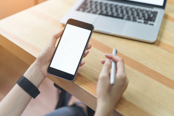 istock woman holding smartphone and using laptop on table in office room on windows with trees and nature background, for graphics display montage. Take your screen to put on advertising. 959560730