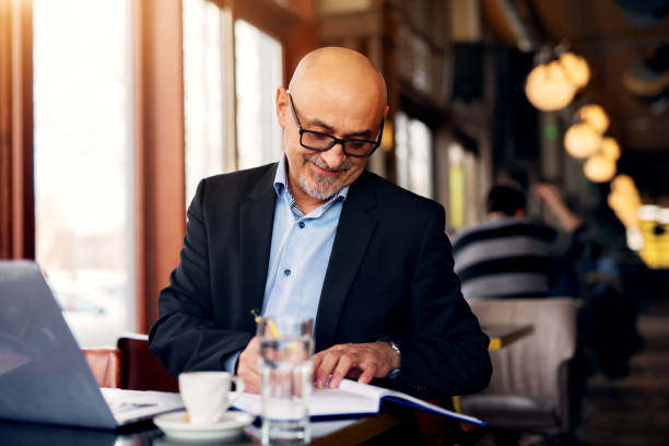 istock Mature professional joyful businessman is using a laptop and drinking coffee while taking notes in his notebook in the coffee shop. 959565488