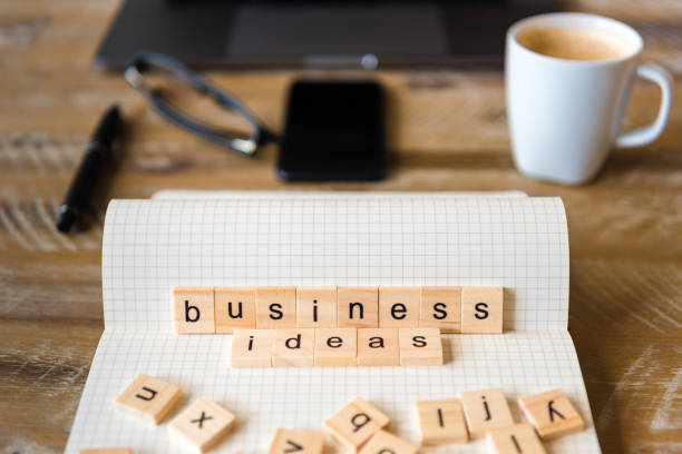 istock Closeup on notebook over wood table background, focus on wooden blocks with letters making Business Ideas words 959565720