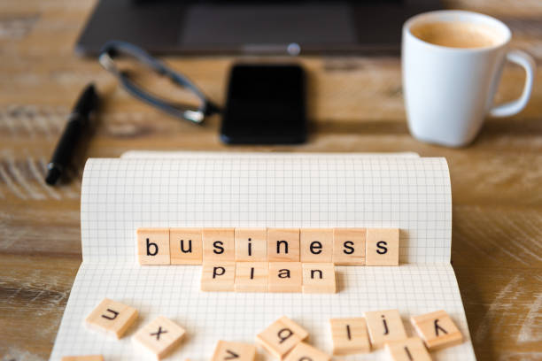istock Closeup on notebook over wood table background, focus on wooden blocks with letters making Business Plan words 959566110