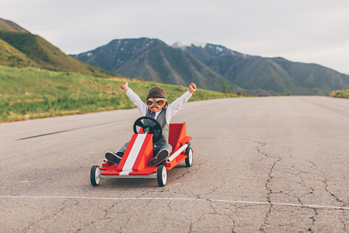 A young business boy dressed in business attire and race goggles in a push cart down a rural road in Utah. This business boy loves racing and competing and working for the success of his business. He gives raised arms at the finish line as he wins.