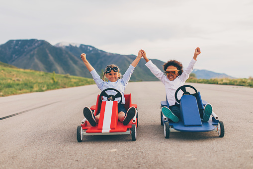 Young business girls dressed in business attire and race goggles in push carts down a rural road in Utah. These business children love racing and competing and working together for the success of their business. They give raised hands in victory at the finish line.