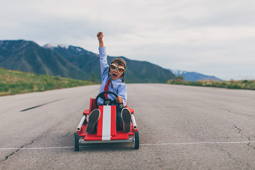 A young business boy dressed in business attire and race goggles races his push cart down a rural road in Utah. He loves racing and competing and working hard for the success of his business. He gives a raised hand in victory at the finish line.