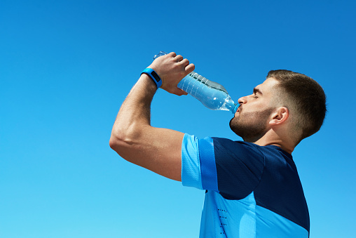 Man Drinking Water After Running. Portrait Of Handsome Athletic Male In Colorful Sportswear Resting After Fitness Workout, Drink Water From Bottle On Blue Sky Background. High Quality Image.