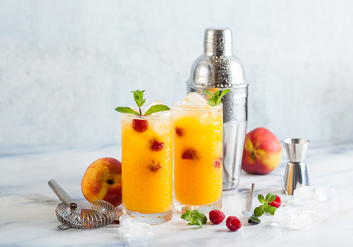 two refreshing drinks from peach juice and raspberries with fresh mint in tall glasses on a marble table with a shaker and bar accessories.