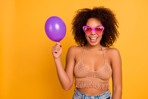 Concept of ease and simplicity. I'm birthday girl today and waiting for presents! Funny relaxed african in summer outfit woman is showing little ultraviolet air balloon, isolated on yellow background