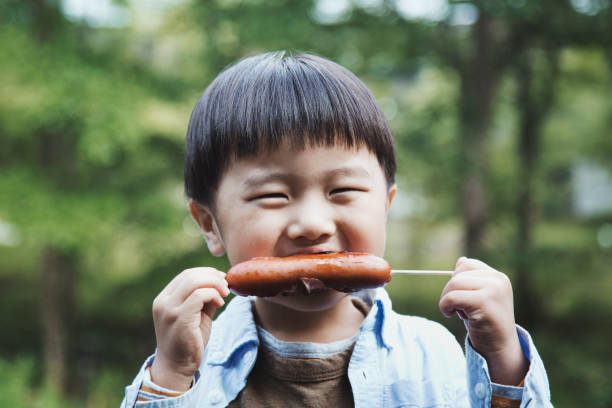 An asian boy eating a sausage cooked for himself at camping.