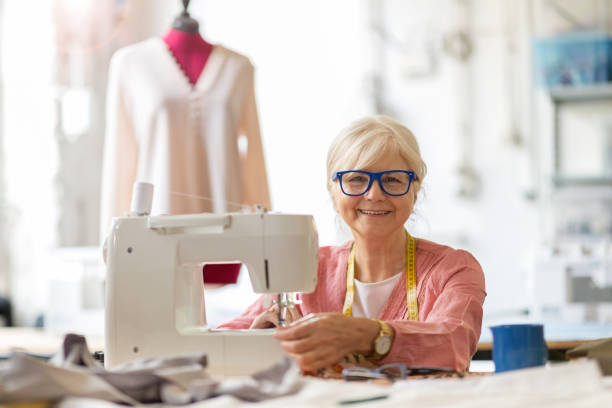 senior fashion designer using a sewing machine in her workshop - manual worker sewing women tailor imagens e fotografias de stock