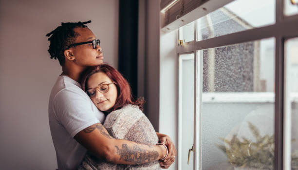 pareja interracial que abrazaba junto a la ventana - parejas abrazadas fotografías e imágenes de stock