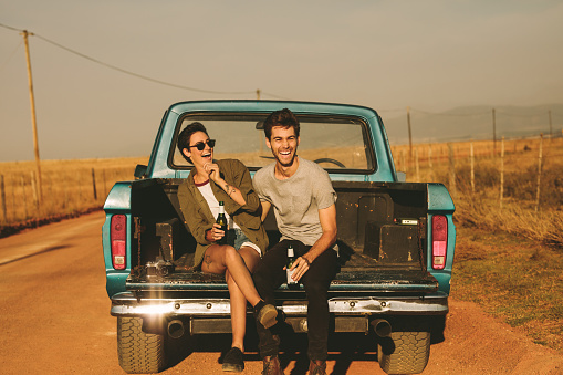 Smiling couple sitting in the back of their pickup truck enjoying the road trip in country side. Man and woman holding a bottle of drink enjoying the road trip.