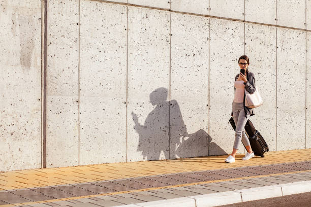 istock Young woman using her smart phone while walking beside the concrete wall and pulling a small wheeled luggage with a briefcase on it 959694880