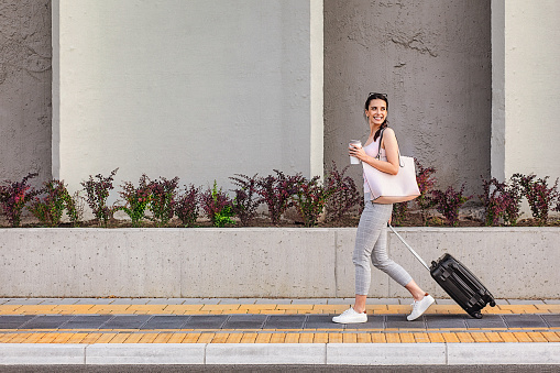 Attractive young woman walking on a sidewalk beside the concrete wall and pulling a small wheeled luggage. She carries a disposable cup of coffee. The shot is executed with available natural light, and the copy space has been left.