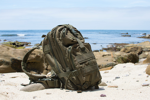Green military backpack sitting on sandy beach with rocky ocean coastline in background on sunny day