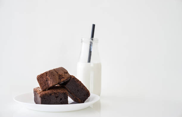 Slice of delicious homemade chocolate brownie served on white plate. Served with glass of milk with green white stripe straw. Taken on a white background. stock photo