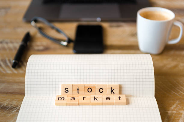 istock Closeup on notebook over wood table background, focus on wooden blocks with letters making Stock Market words 959721088